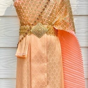 May include: A peach-coloured traditional Thai dress with gold embroidery and a matching sash. The dress has a pleated skirt and a sheer overlay. The dress is decorated with a flower crown.