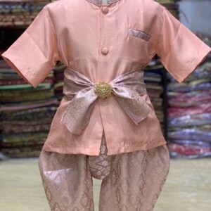 May include: A peach-colored traditional Thai child's outfit. The set includes a short-sleeved shirt with a mandarin collar, matching pants, and a decorative sash with a gold-colored embellishment. The outfit is displayed on a mannequin.