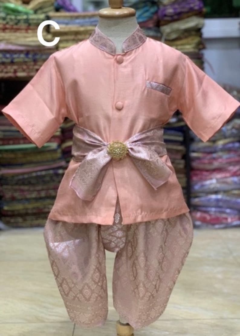 May include: A peach-colored traditional Thai child's outfit. The set includes a short-sleeved shirt with a mandarin collar, matching pants, and a decorative sash with a gold-colored embellishment. The outfit is displayed on a mannequin.