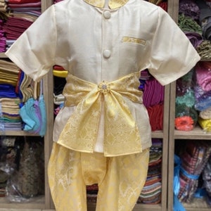 May include: A cream-colored traditional outfit for a child, featuring a short-sleeved shirt with gold trim and buttons, paired with matching pants. A decorative gold sash with a jeweled clasp accents the waist. The outfit is displayed on a mannequin.