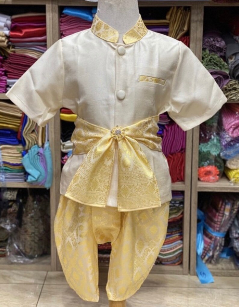 May include: A cream-colored traditional outfit for a child, featuring a short-sleeved shirt with gold trim and buttons, paired with matching pants. A decorative gold sash with a jeweled clasp accents the waist. The outfit is displayed on a mannequin.