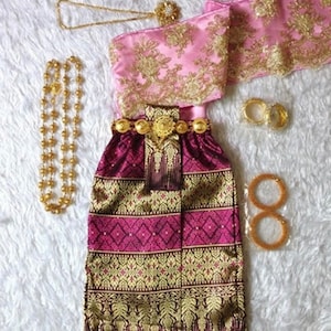May include: A traditional Thai outfit featuring a burgundy skirt with gold embroidery, a pink sash with gold embroidery, and gold jewellery.