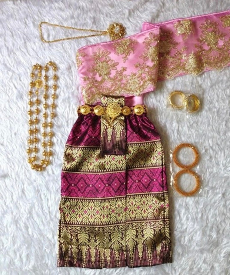 May include: A traditional Thai outfit featuring a burgundy skirt with gold embroidery, a pink sash with gold embroidery, and gold jewellery.