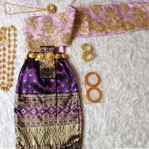 May include: A purple and gold traditional Thai dress with a matching sash and jewellery. The dress has a patterned skirt and a top with gold embroidery. The jewellery includes a necklace, bracelets, and a belt.