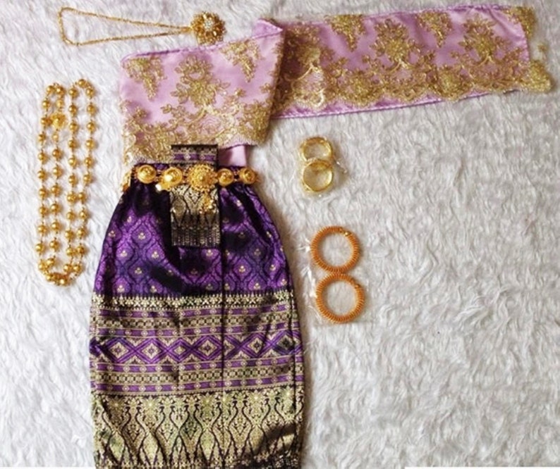 May include: A purple and gold traditional Thai dress with a matching sash and jewellery. The dress has a patterned skirt and a top with gold embroidery. The jewellery includes a necklace, bracelets, and a belt.