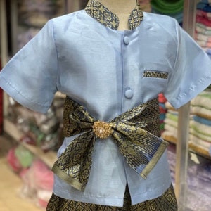 May include: A light blue traditional outfit for a child. The short-sleeved shirt has a mandarin collar and is buttoned down the front. The outfit includes patterned pants and a decorative sash with a gold-colored brooch.