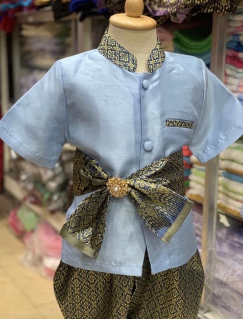 May include: A light blue traditional outfit for a child. The short-sleeved shirt has a mandarin collar and is buttoned down the front. The outfit includes patterned pants and a decorative sash with a gold-colored brooch.