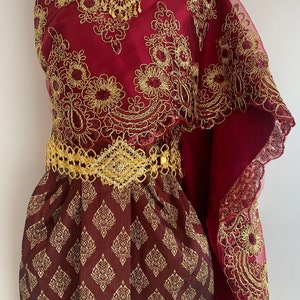 May include: A maroon and gold traditional Thai dress with intricate embroidery and a gold belt. The dress has a flowing, sheer overlay with a scalloped edge.