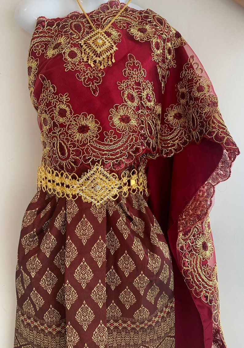 May include: A maroon and gold traditional Thai dress with intricate embroidery and a gold belt. The dress has a flowing, sheer overlay with a scalloped edge.