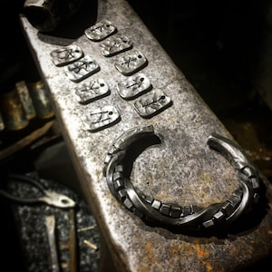 May include: A collection of handcrafted silver jewelry pieces, including several small rectangular tags with etched symbols and a textured cuff bracelet. The items are displayed on a metal surface, showcasing a rustic, artisan aesthetic.