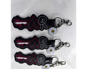 Könnte beinhalten: Drei schwarze Schlüsselanhänger mit gestickten Motiven. Jeder Anhänger zeigt eine bordeaux-weiß gefärbte Flaggenform, ein schwarzes Herz mit roten Nähten und eine Handgeste für Frieden. Ein kleiner weißer Gänseblümchen-Anhänger hängt an jedem Ring.
