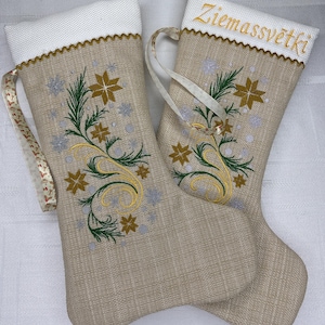 May include: Two beige Christmas stockings with white tops and gold trim. Each stocking features embroidered gold, green, and silver floral designs. One stocking has the word "Ziemassvētki" embroidered in gold.