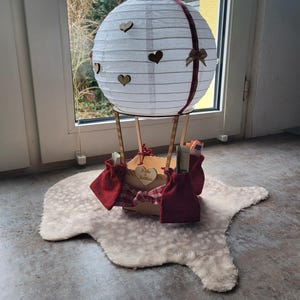 May include: A white paper lantern hot air balloon decoration featuring small wooden heart cutouts, a dark red ribbon, and a wooden basket base. The basket is adorned with red fabric pouches and a small wooden heart engraved with the text Katja & Matthias.
