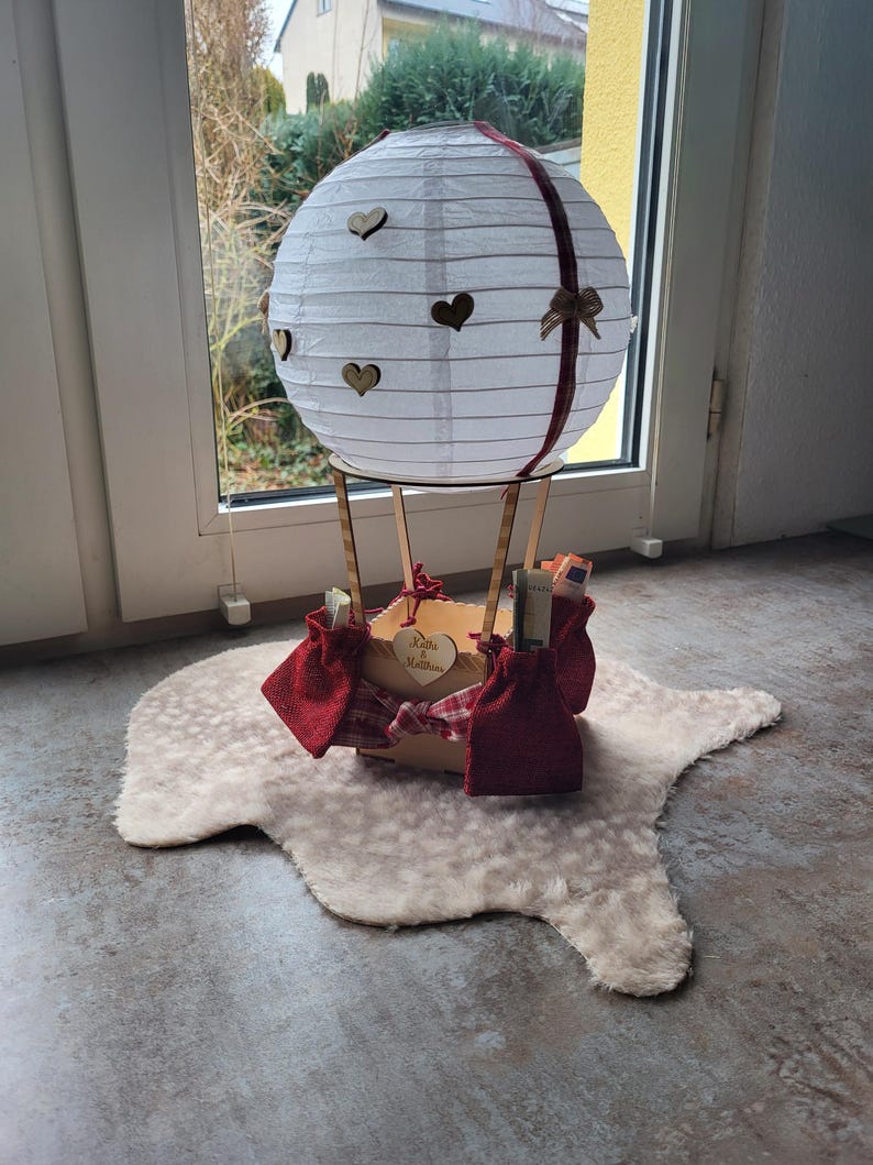 May include: A white paper lantern hot air balloon decoration featuring small wooden heart cutouts, a dark red ribbon, and a wooden basket base. The basket is adorned with red fabric pouches and a small wooden heart engraved with the text Katja & Matthias.