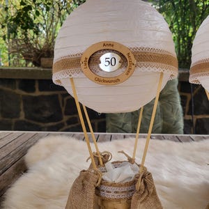 Könnte beinhalten: Dekorative Heißluftballon-förmige Tischdekoration. Die weiße Papierlaterne ist mit einem Jutegürtel, Spitzentrimm und einem Holzring mit der Zahl 50 und dem Namen "Anne" verziert. Der Korb besteht aus gewebtem Material und Jute.
