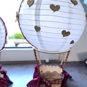 May include: A decorative hot air balloon ornament featuring a white paper lantern globe adorned with small wooden heart cutouts. The basket is made of light-colored wood, accented with dark red fabric ribbons and a small heart-shaped wooden plaque.