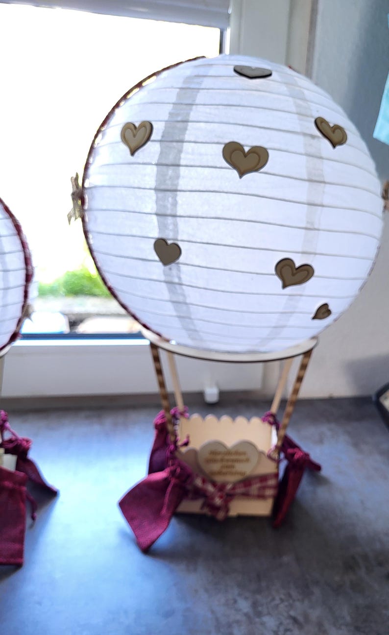 May include: A decorative hot air balloon ornament featuring a white paper lantern globe adorned with small wooden heart cutouts. The basket is made of light-colored wood, accented with dark red fabric ribbons and a small heart-shaped wooden plaque.