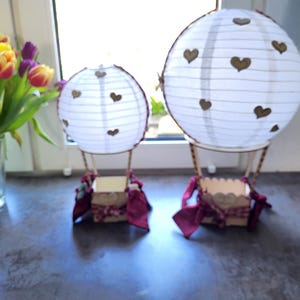 May include: Two decorative hot air balloon models featuring white paper lanterns with small brown heart cutouts. Each balloon has a wooden basket base adorned with dark red fabric ribbons and bows, creating a pair of matching tabletop ornaments.