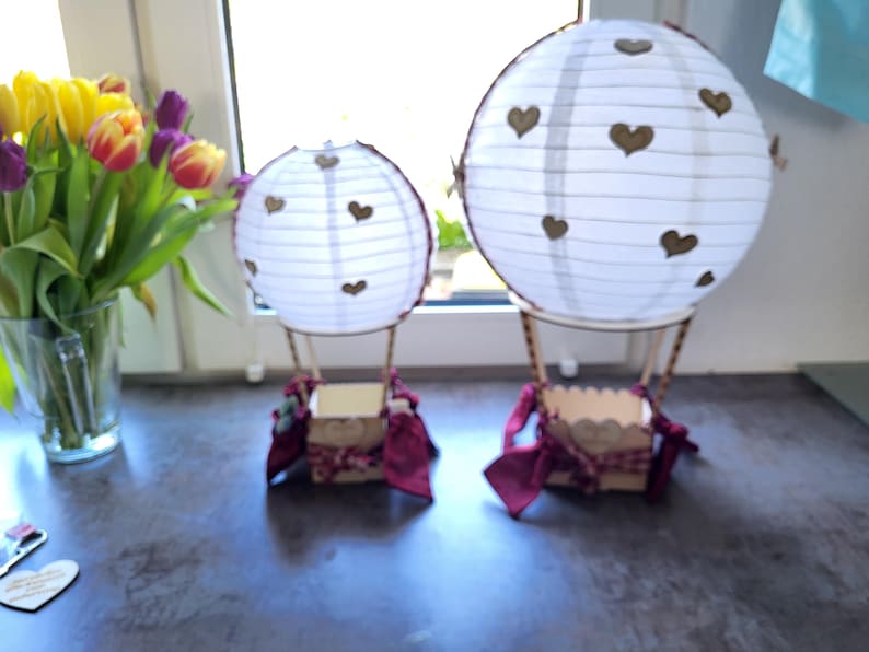 May include: Two decorative hot air balloon models featuring white paper lanterns with small brown heart cutouts. Each balloon has a wooden basket base adorned with dark red fabric ribbons and bows, creating a pair of matching tabletop ornaments.