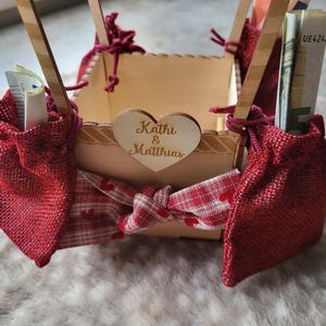 May include: A square wooden box with a heart-shaped cutout featuring the text Kathi & Matthias. The box is decorated with red burlap pouches, a red and white checkered ribbon with heart patterns, and wooden supports holding rolled-up currency.