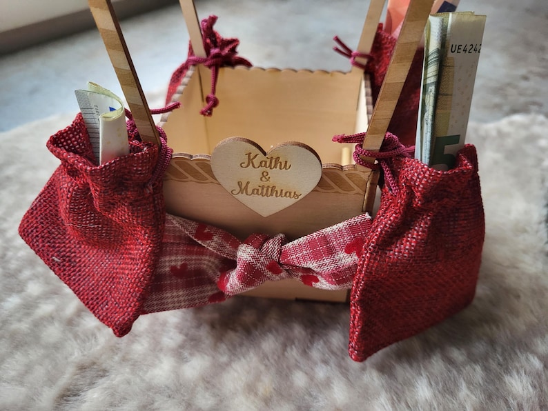 May include: A square wooden box with a heart-shaped cutout featuring the text Kathi & Matthias. The box is decorated with red burlap pouches, a red and white checkered ribbon with heart patterns, and wooden supports holding rolled-up currency.