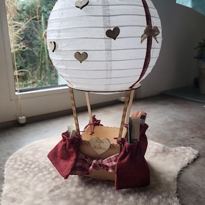 May include: A white paper lantern hot air balloon decoration featuring small wooden heart cutouts, a red plaid ribbon, and a wooden basket base. The basket is adorned with red burlap pouches and a heart-shaped wooden tag engraved with Kathi & Matthias.
