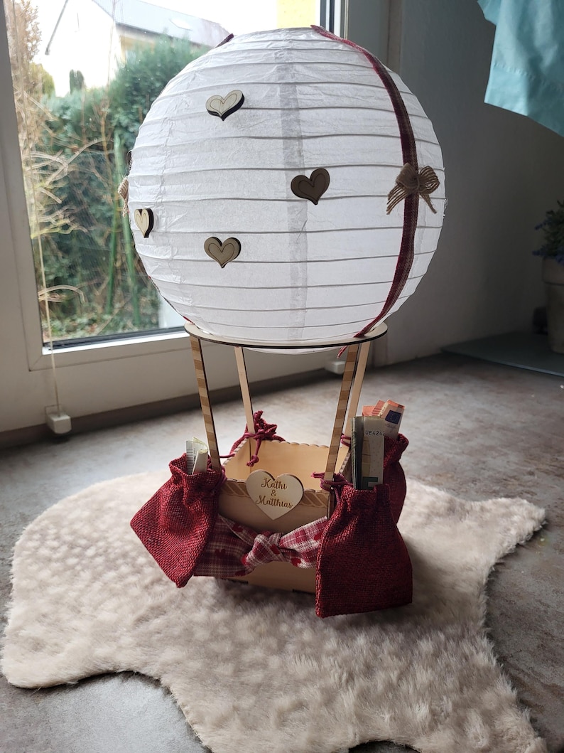 May include: A white paper lantern hot air balloon decoration featuring small wooden heart cutouts, a red plaid ribbon, and a wooden basket base. The basket is adorned with red burlap pouches and a heart-shaped wooden tag engraved with Kathi & Matthias.