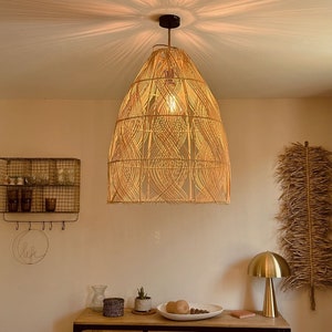 Könnte beinhalten: Eine große, gewebte, hellbraune Pendelleuchte mit geometrischem Muster hängt von der Decke. Die Lampe ist eingeschaltet und strahlt ein warmes Licht in den Raum. Die Lampe befindet sich in einem Raum mit einem Holztisch, einer kleinen Pflanze und einer goldenen Tischlampe.