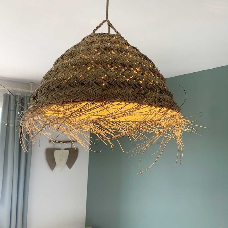 May include: A woven wicker pendant light with a fringed bottom. The light is hanging from a white ceiling and is illuminating the room with a warm glow.