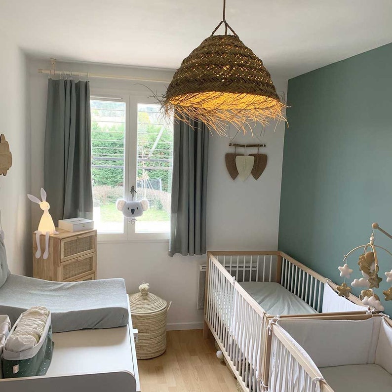 May include: A nursery with two white cribs, a changing table, and a woven light fixture. The walls are painted a light green and the curtains are a light gray. There is a white stuffed animal hanging from the window.