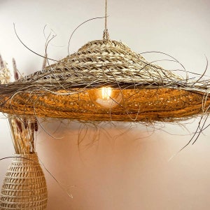 May include: A woven straw pendant light fixture with a wide, flat, circular shade. The light is turned on and illuminates the room with a warm, yellow glow.
