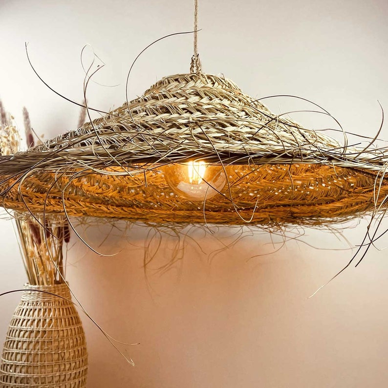 May include: A woven straw pendant light fixture with a wide, flat, circular shade. The light is turned on and illuminates the room with a warm, yellow glow.