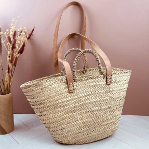 Peut inclure: Un sac fourre-tout en paille tressée avec des poignées en cuir beige et une longue bandoulière. Le sac est de couleur marron clair naturel et a un aspect texturé. Les poignées sont fixées avec des coutures visibles.