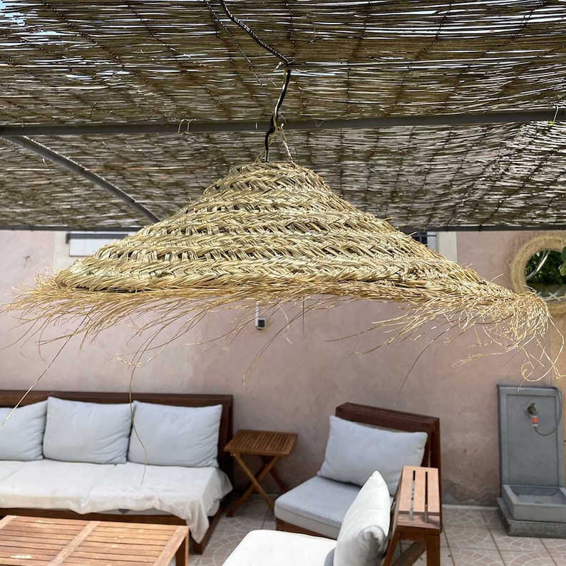 May include: A large, round, woven straw lampshade hangs from a hook. The lampshade is light brown and has a fringe around the bottom edge.