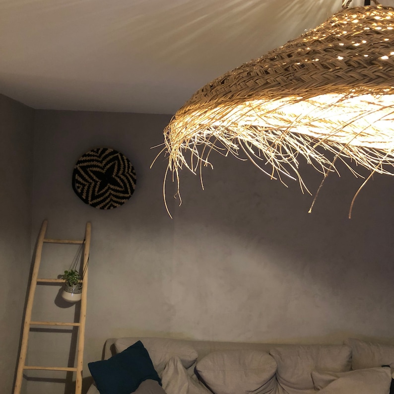 May include: A large, round, woven, straw lampshade hangs from the ceiling in a room with a light grey wall. The lampshade is lit and casts a warm glow on the wall. A small, round, woven wall hanging with a black and white star design is on the wall.