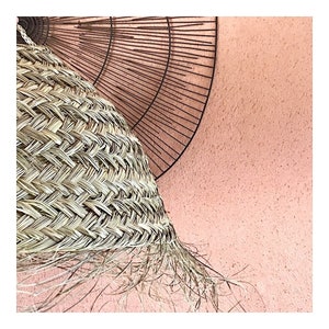 May include: A woven wicker lampshade with a fringe hanging from the bottom. The lampshade is against a light pink wall.