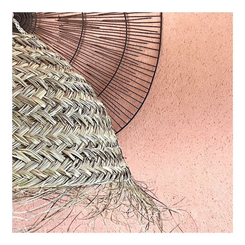 May include: A woven wicker lampshade with a fringe hanging from the bottom. The lampshade is against a light pink wall.
