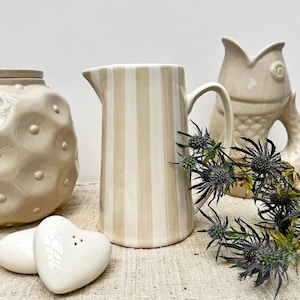 Può includere: Un'anfora in ceramica a righe con un design beige e bianco. L'anfora è appoggiata su una tovaglia di lino beige con un bouquet di cardi secchi.