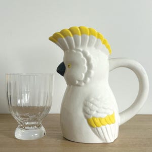 Peut inclure: Un pichet en céramique blanche en forme de cacatoès avec des accents jaunes. Le pichet a un bec verseur et une anse. Un verre transparent à nervures verticales est à côté du pichet.