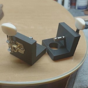 May include: Two black guitar tuning machines with cream-colored knobs and gold-colored gears. The tuning machines are positioned on a wooden surface, likely a guitar body, showcasing their design and functionality.