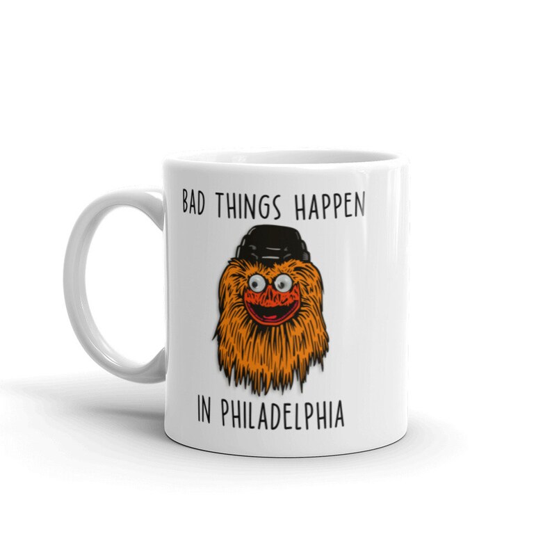May include: White ceramic mug with a cartoon orange and brown furry creature wearing a black hat. The text "Bad things happen in Philadelphia" is printed on the mug.