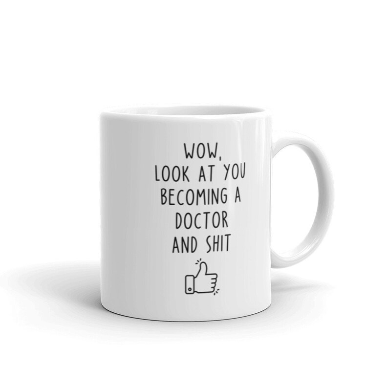 May include: White ceramic coffee mug with black text that reads "WOW, LOOK AT YOU BECOMING A DOCTOR AND SHIT" with a thumbs up emoji.