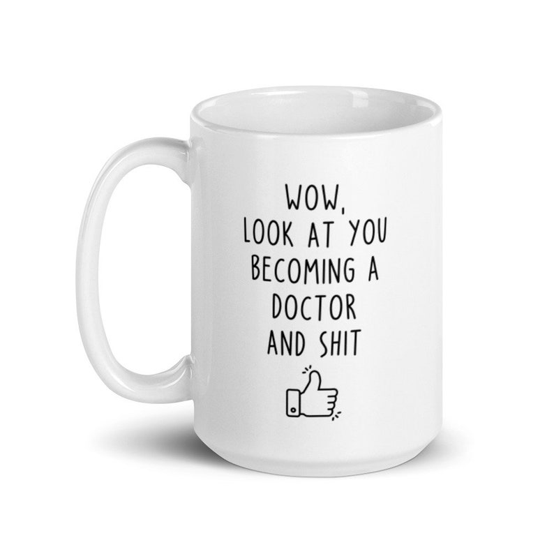 May include: White ceramic coffee mug with black text that reads "WOW, LOOK AT YOU BECOMING A DOCTOR AND SHIT" with a thumbs up emoji.