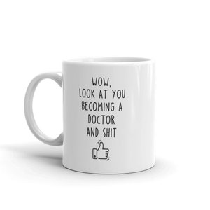 May include: White ceramic coffee mug with black text that reads "WOW, LOOK AT YOU BECOMING A DOCTOR AND SHIT" with a thumbs up emoji.