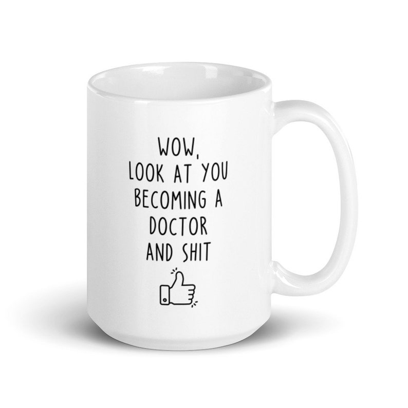 May include: White ceramic coffee mug with a black inscription that reads "WOW, LOOK AT YOU BECOMING A DOCTOR AND SHIT" with a thumbs up emoji.