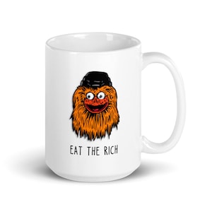 May include: White ceramic mug with a cartoon illustration of a furry orange monster wearing a black hat. The text "EAT THE RICH" is printed below the illustration.