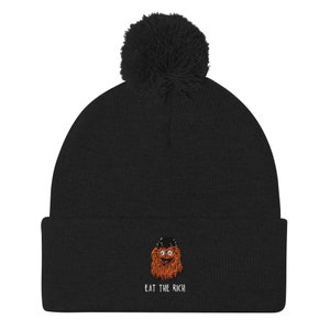 May include: Black knitted beanie with a fluffy pom pom on top. The beanie features an embroidered image of a cartoon character with a red beard and the text "EAT THE RICH".