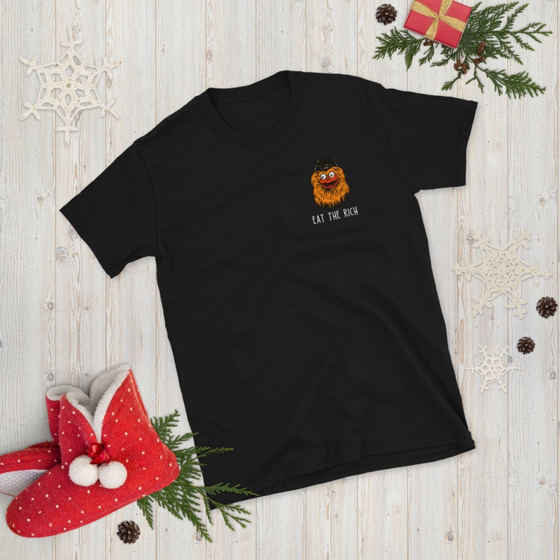 Op de afbeelding: Zwart T-shirt met een cartoon afbeelding van een harig oranje monster met een hoed en de tekst "EAT THE RICH" gedrukt op de voorkant.