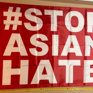 May include: A red flag with the words "#STOP ASIAN HATE" in large, white capital letters. The flag is rectangular and appears to be made of a lightweight fabric. It is hanging against a neutral background.