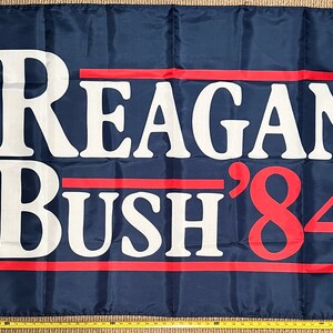 May include: A blue political campaign flag with white text that reads "Reagan Bush '84" in a bold, sans-serif font. Two red stripes are above and below the text.